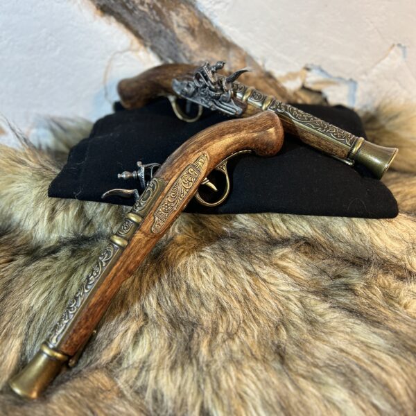 Två dekorativa flintlåspistoler i mörkt trä med mässingsdetaljer, liggande på svart tyg och fäll mot rustik vägg