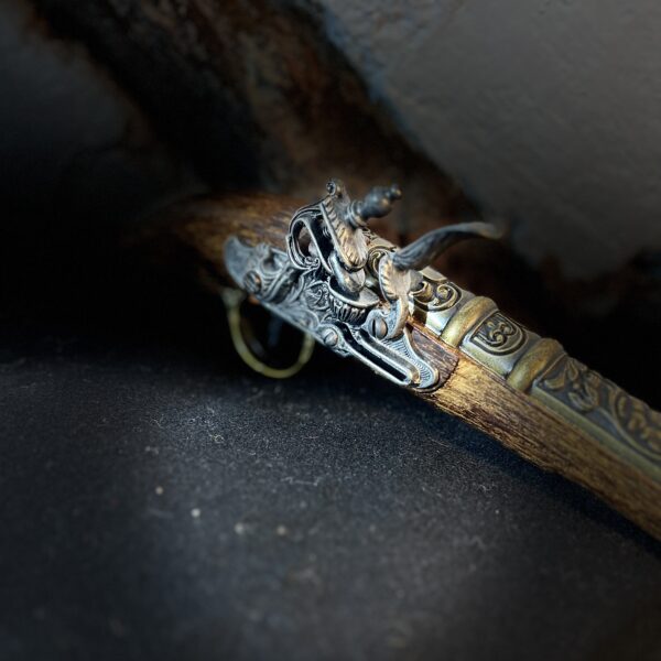Detalj från en dekorativ flintlåspistol i mörkt trä med mässingsdetaljer, liggande på svart tyg och fäll mot rustik vägg