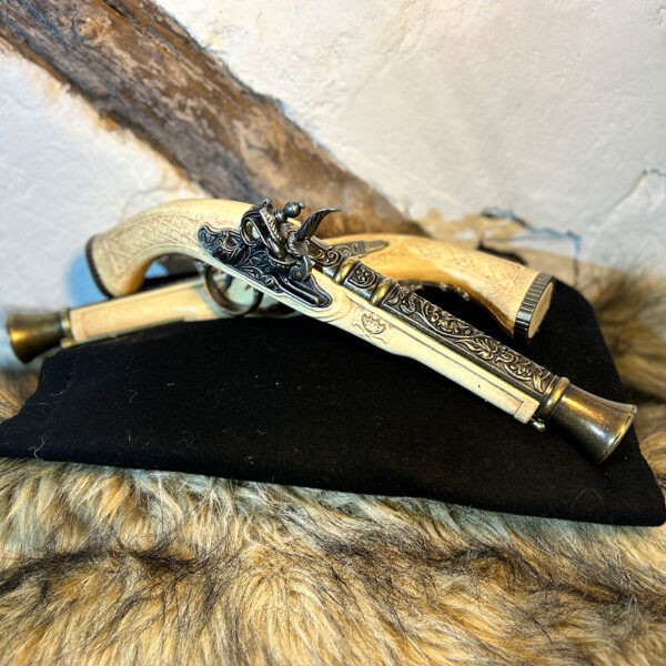 Två dekorativa flintlåspistoler i elfenbensimitation med detaljerade metallbeslag, liggande på svart tyg och fäll