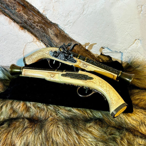 Två dekorativa flintlåspistoler i elfenbensimitation med detaljerade metallbeslag, liggande på svart tyg och fäll